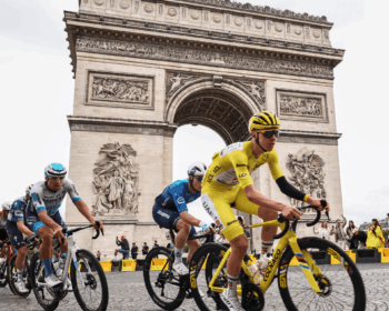 Group of pro cyclists racing past the Arc de Triomphe in Paris, yellow-clad rider leading on a cobblestone street